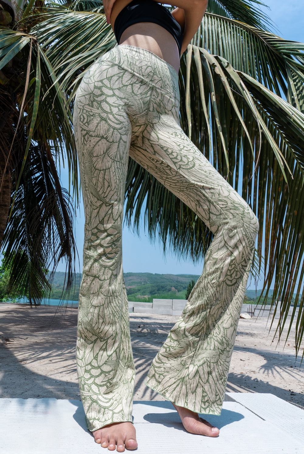 Model wearing Funky Simplicity Flares in Cream Olive Feather print made from cotton lycra - with green palm trees and a river on the background - shown from the front