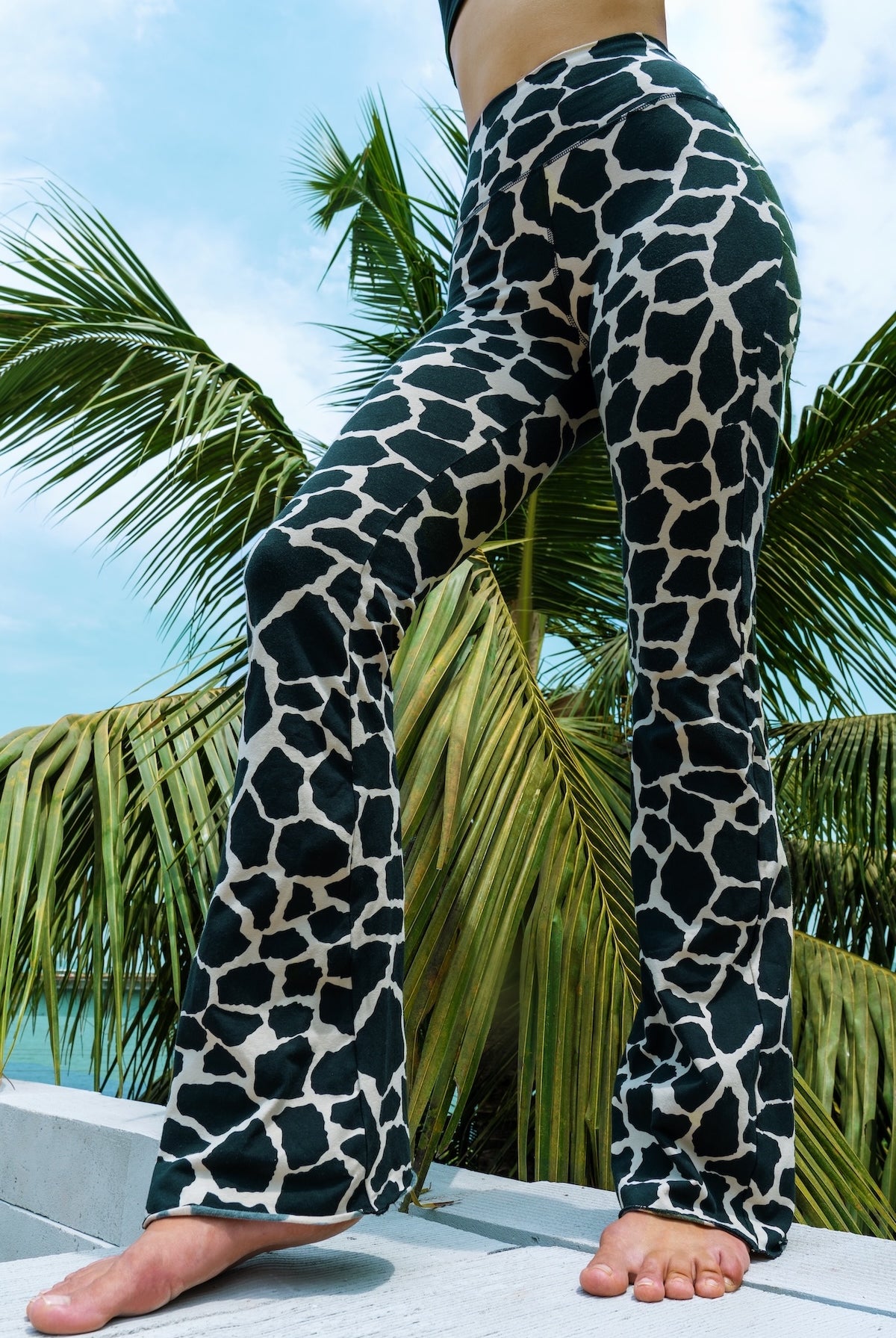 Woman wearing cotton lycra Flares in Cream Black Dutch Cow print from Funky Simplicity shown from the front with leaves of a palmtree in the background with a cloudy blue sky