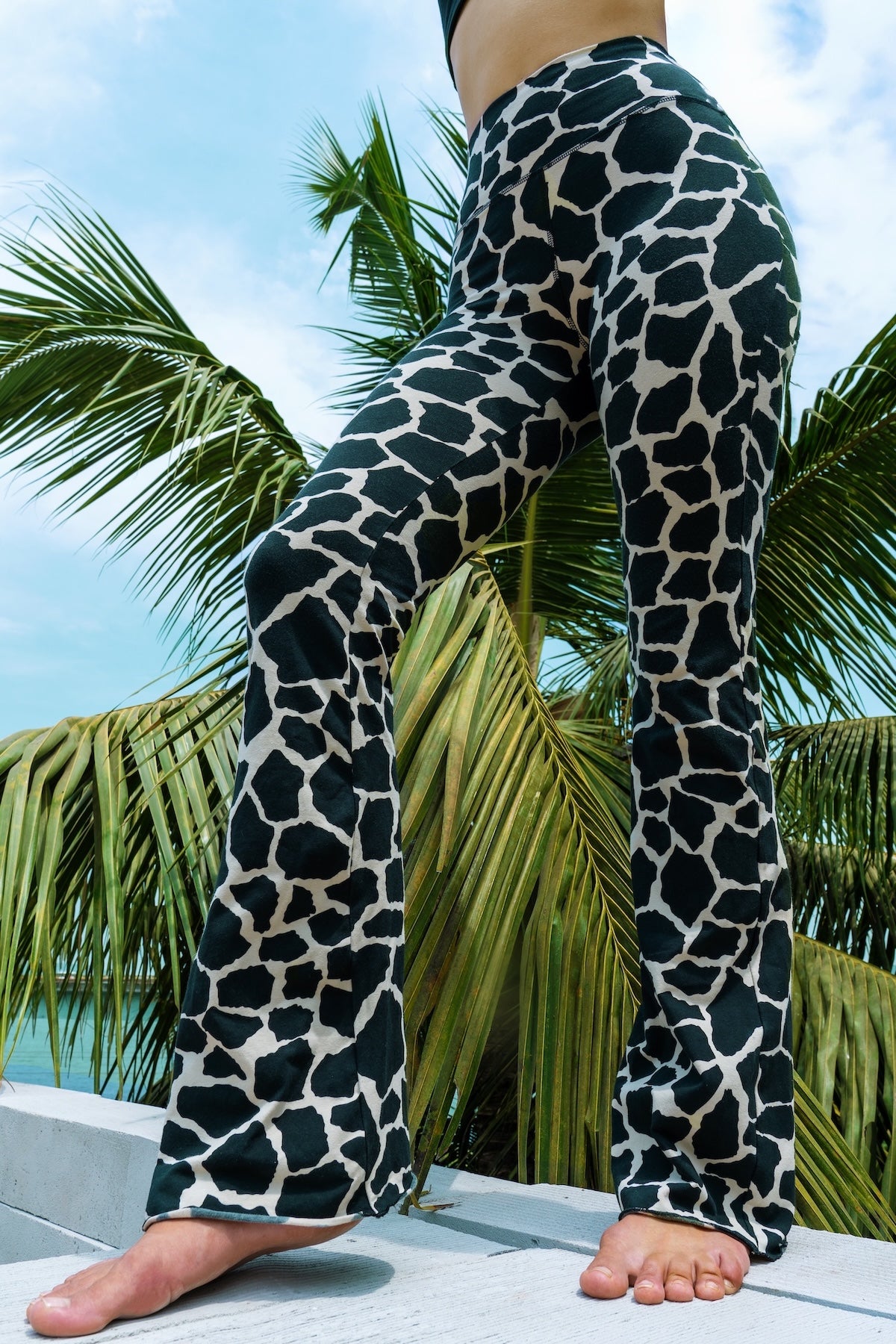 Woman wearing cotton lycra Flares in Cream Black Dutch Cow print from Funky Simplicity shown from the front with leaves of a palmtree in the background with a cloudy blue sky