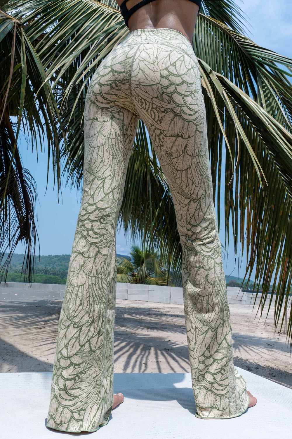 Model wearing Funky Simplicity Flares in Cream Olive Feather print made from cotton lycra - with green palm trees and a river on the background - shown from the back