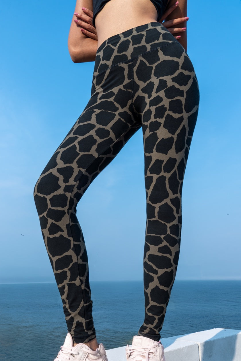 Woman wearing Funky Simplicity Giraffe Leggings in the colour Olive Black shown from the front with an ocean and blue sky on the background