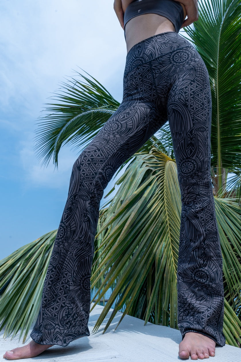 Model wearing Funky Simplicity tribal-print flared leggings in grey black color shown from the front with green palm trees and a blue sky with some clouds on the background.